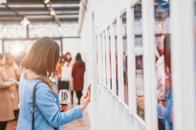 A woman in an art gallery takes a picture of an art piece title card while holding a glass of red wine